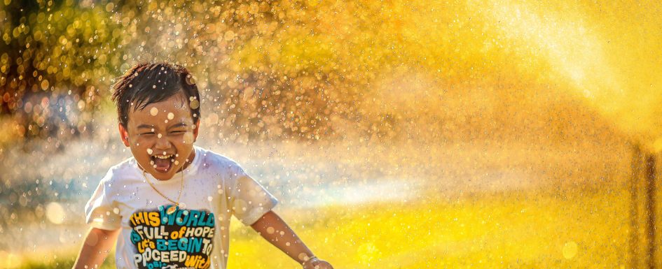 A young child wearing a T-shirt that reads, "This world is full of hope" plays in a sprinkler. The water is catching the afternoon sunlight and glowing yellow.