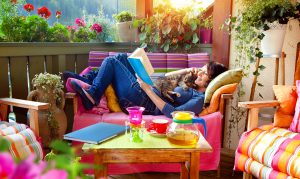 A white woman is curled up on a balcony chair, reading a book and petting a sleepy cat. She is surrounded by brightly-colored flowers and furniture.